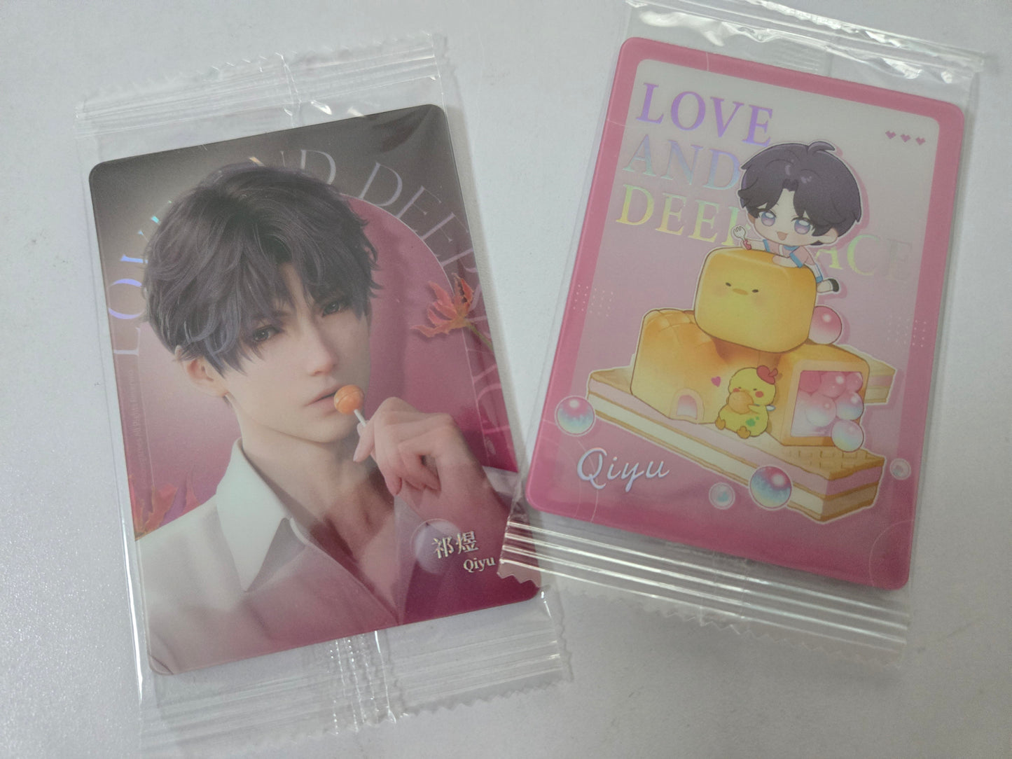 Two sealed collectible cards in clear plastic packaging on a light gray surface. One card shows Rafayel in a white shirt against a pink background, and the other shows a pink card with the text 'LOVE AND DEEPER Qiyu' and a cartoon character above stacked yellow blocks.