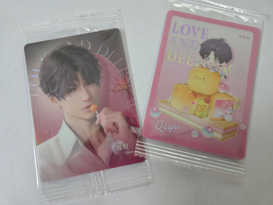 Two sealed collectible cards in clear plastic packaging on a light gray surface. One card shows Rafayel in a white shirt against a pink background, and the other shows a pink card with the text 'LOVE AND DEEPER Qiyu' and a cartoon character above stacked yellow blocks.