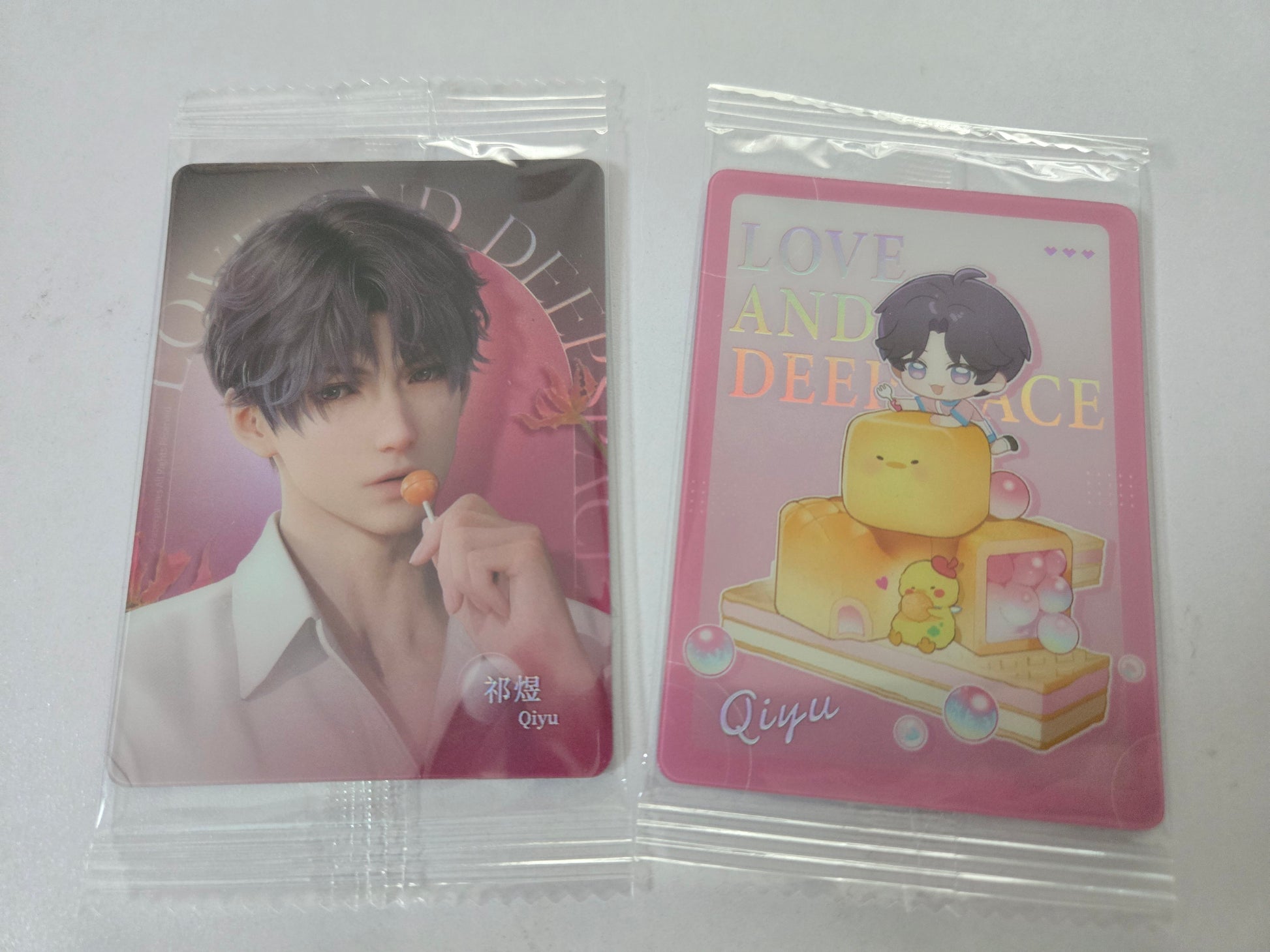 Two sealed collectible cards in clear plastic sleeves on a white background. The left card shows a portrait of Rafayel with purple hair holding an orange lollipop, with pink Love and Deepspace text.